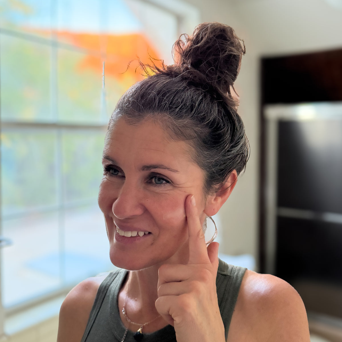 A smiling woman with her hair in a messy bun rubs tallow balm on cheek, wearing a dark olive green tank top in a brightly lit indoor setting.