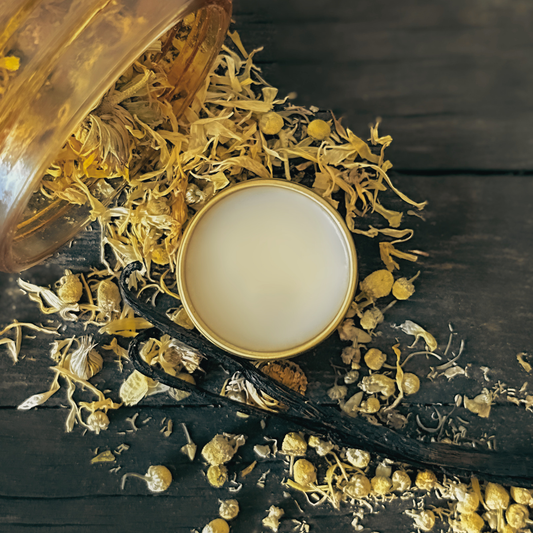 Vanilla Petal balm with dried chamomile, calendula, and vanilla bean scattered from a jar around it on a dark wooden surface