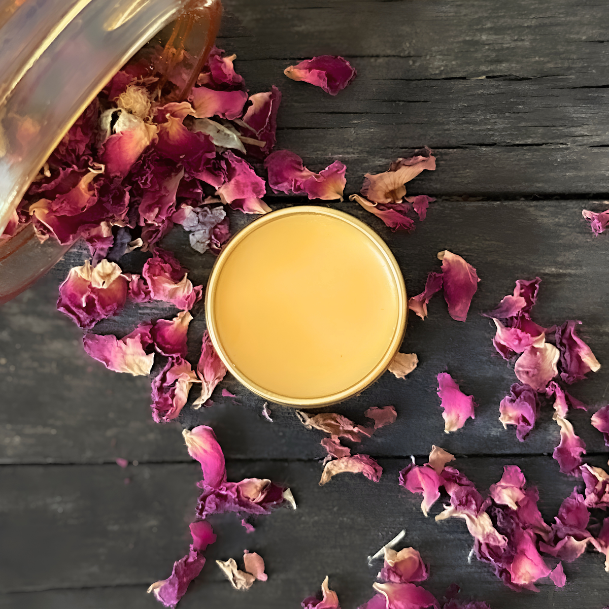 Strawberry Bloom balm with dried red rose petals scattered from a jar around it on a dark wooden surface