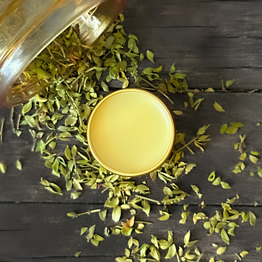 Mojave Rain balm with dried creosote scattered from a jar around it on a dark wooden surface